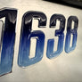 Metallic badge with numbers '1638 JB' on a reflective surface